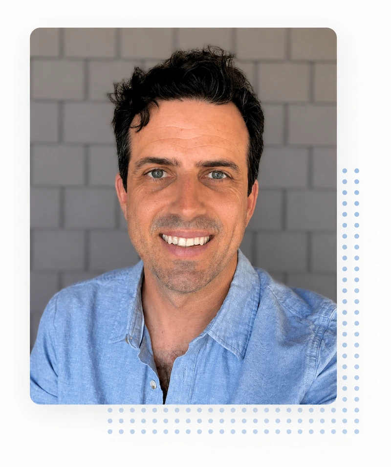 Man with dark hair and blue eyes smiles at the camera, wearing a light blue shirt, standing in front of a gray brick wall.